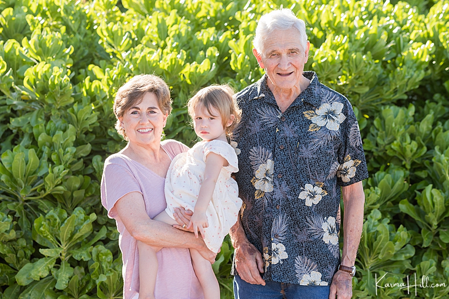 grandparents holding granddaughter with green bushes in background 