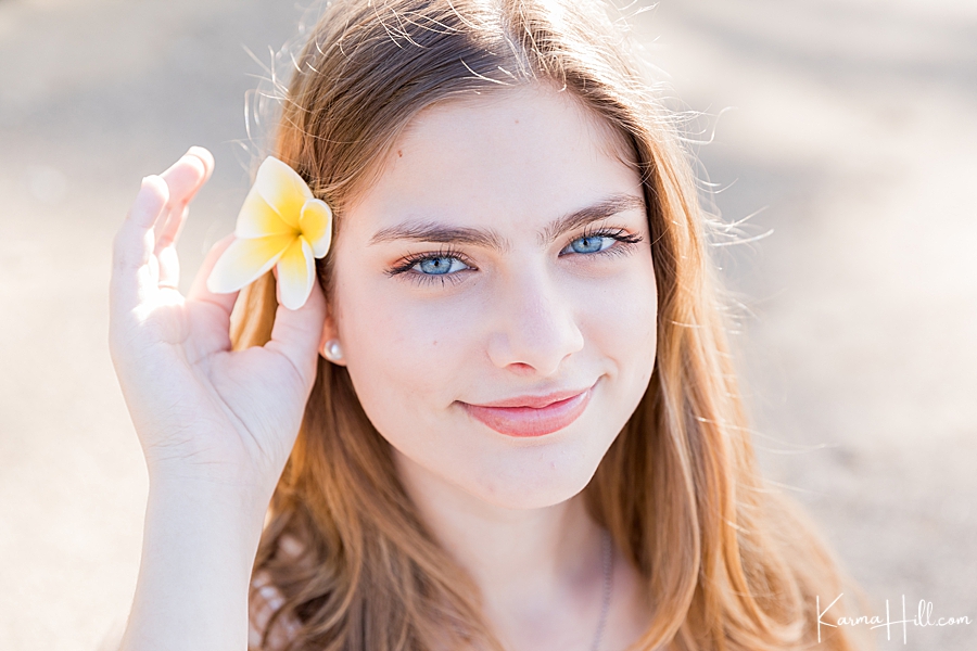 young woman with red hair and blue eyes holds plumeria flower in her ear on hawaii beach 