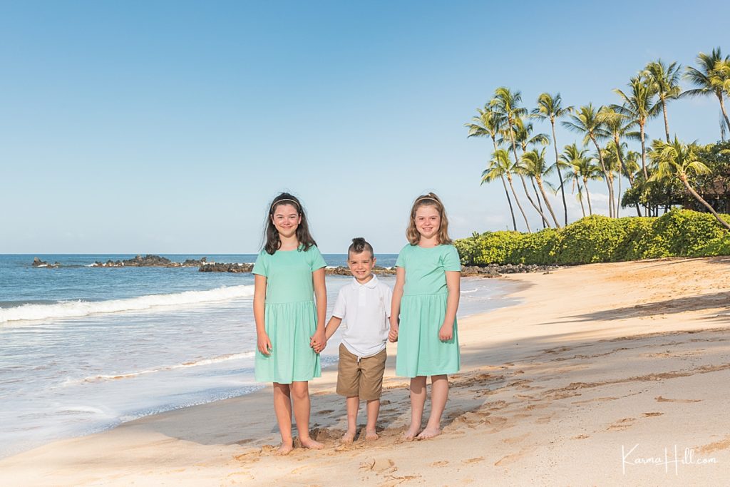 three children hold hands wearing aqua and white on a hawaii beach with palm trees in the background 