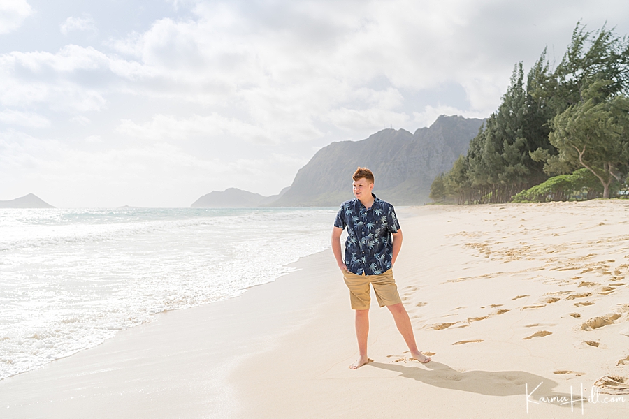 young man with red hair stands on an oahu beach wearing aloha shirt and beige shorts 