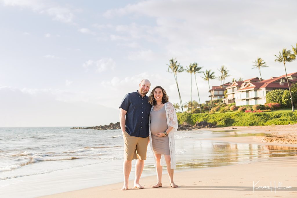 couple stand on maui beach with woman holding her pregnant belly 