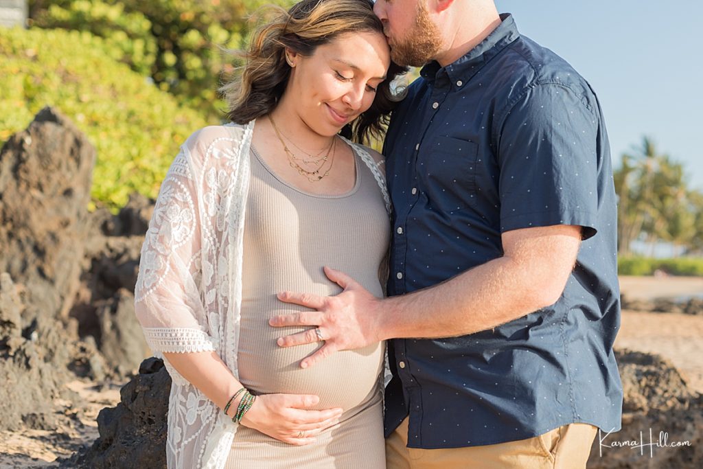 man places hand on woman's pregnant belly and kisses her forehead on a maui beach 