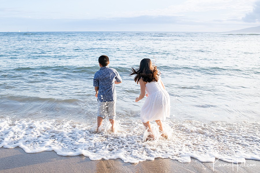 brother and sister leap in ocean waves wearing a white dress and aloha shirt 