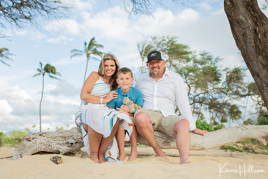 family of three sit on a driftwood log in a maui beach with palm trees in the background 