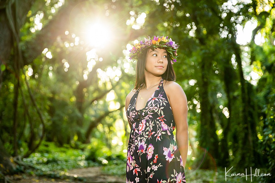 a young woman wearing a flower crown and floral dress stands on the honolua jungle path 