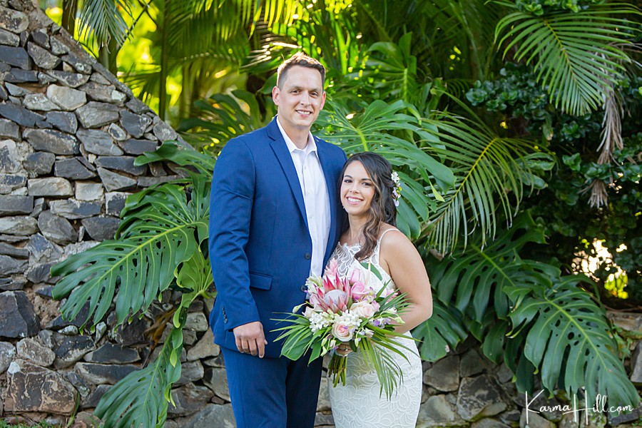 bride and groom stand in front of monstera leaves and a rock wall in hawaii 