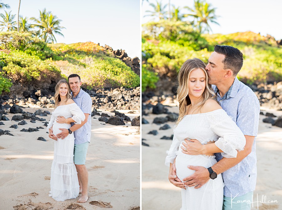 expecting mother wearing a white dress is held by her husband with his hands on her belly on the shore of a maui beach 