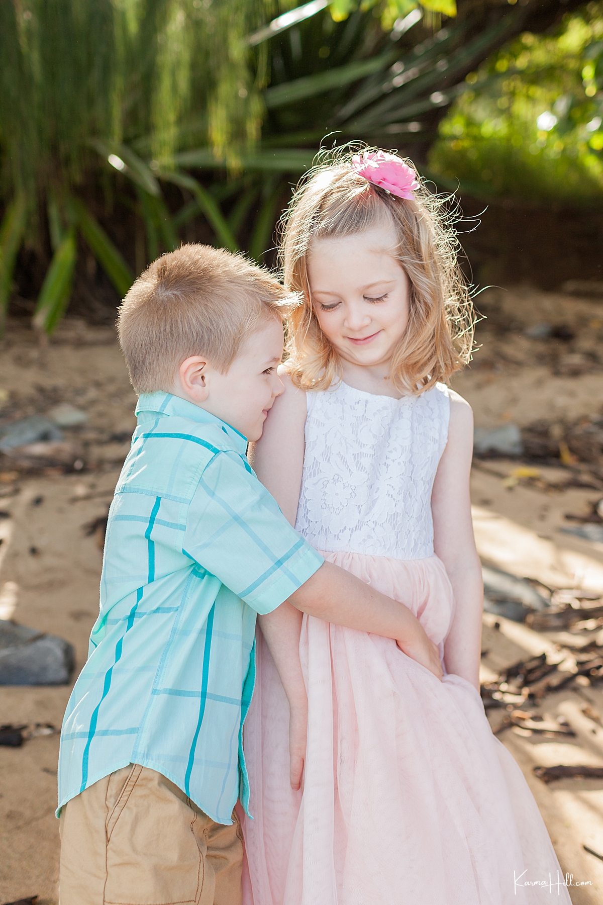 little brother hugs his older sister as she smiles down at him. Brother wears a blue collared shirt and sister is in a lace flower dress with coral tulle