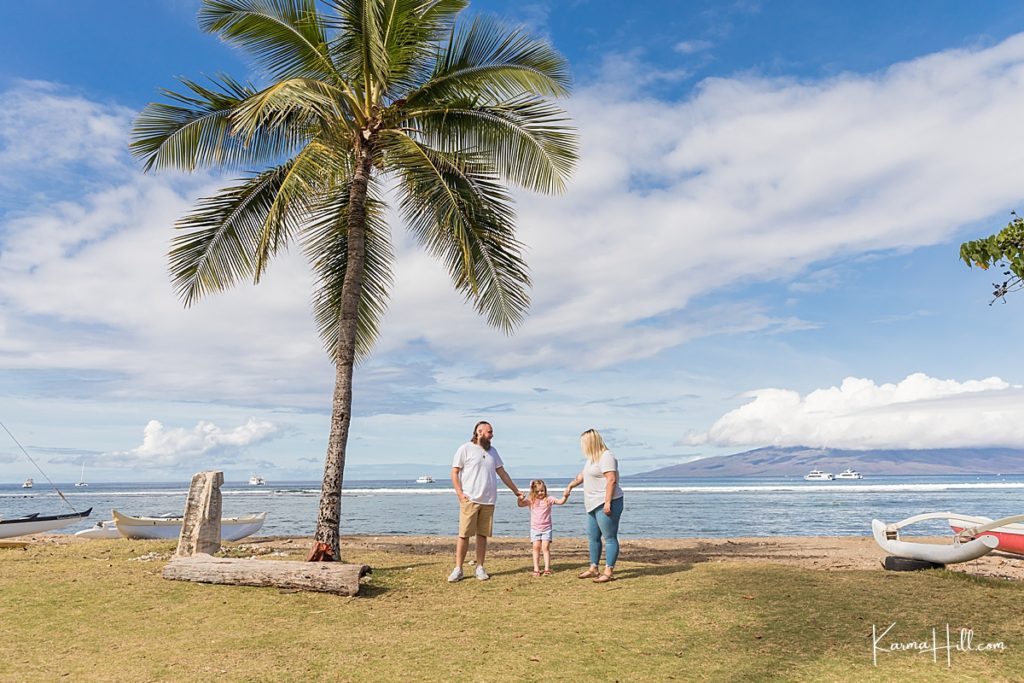 family of three stands beneath palm tree with boats and the ocean in the distance 