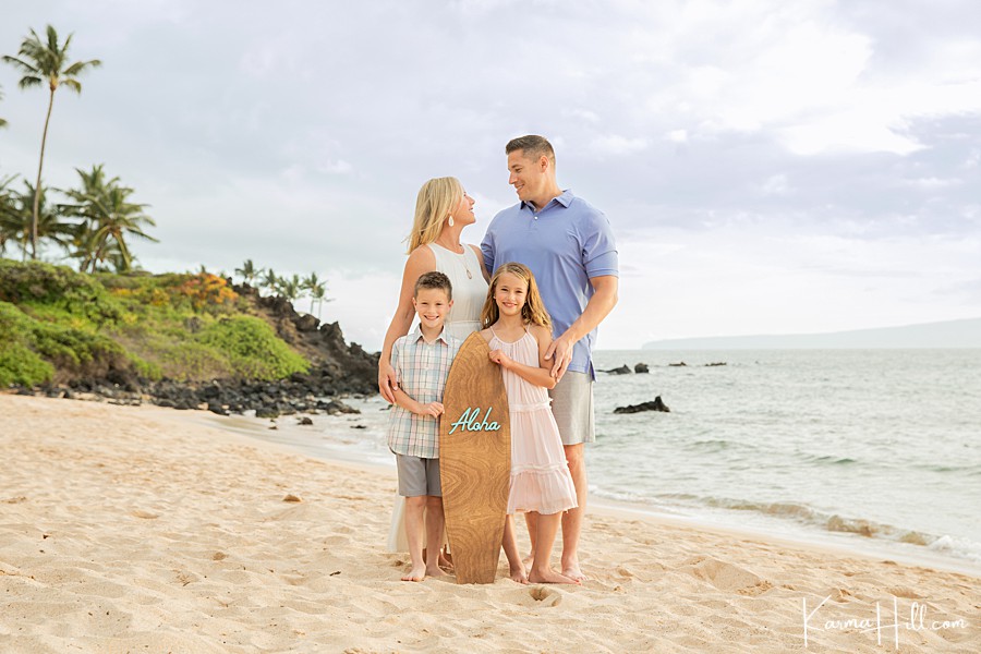 family of four with mom dad brother and sister pose with an aloha surfboard on hawaii beach 