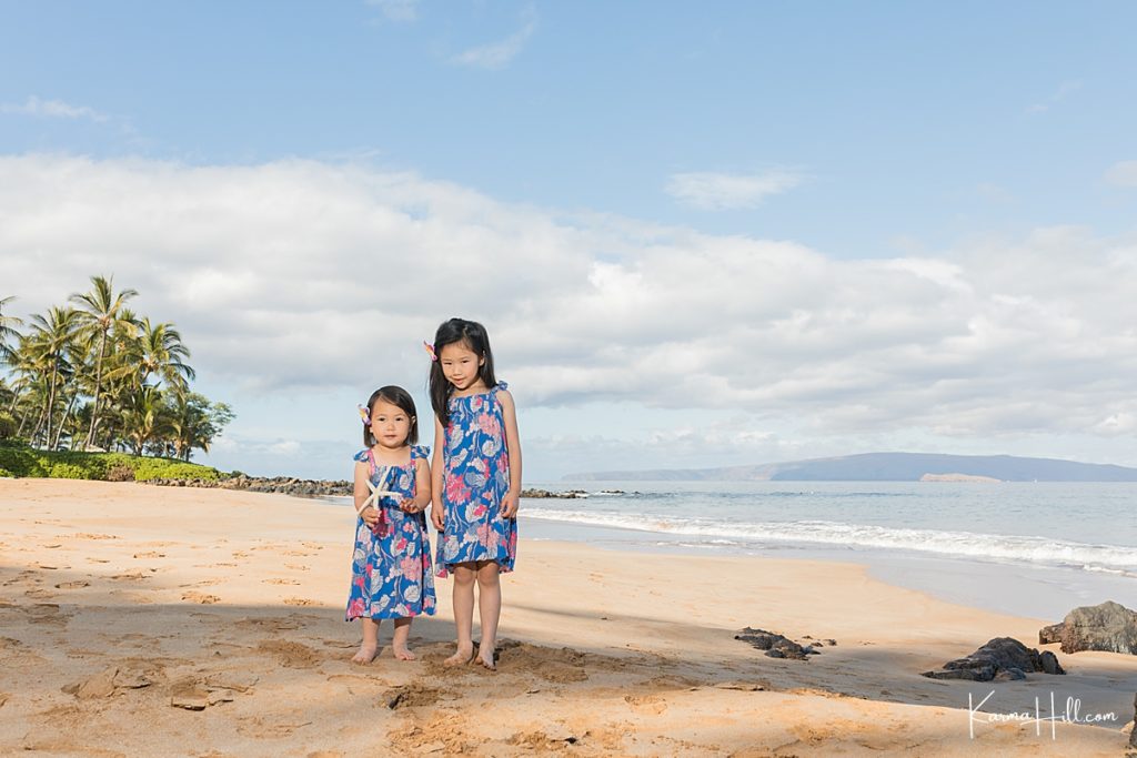 two little girls stand on a tropical beach with palm trees in the background holding a starfish 