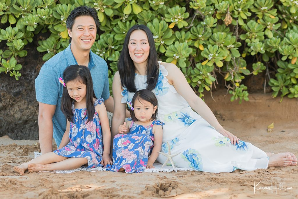 family of four sit on the sand with greenery in the background 