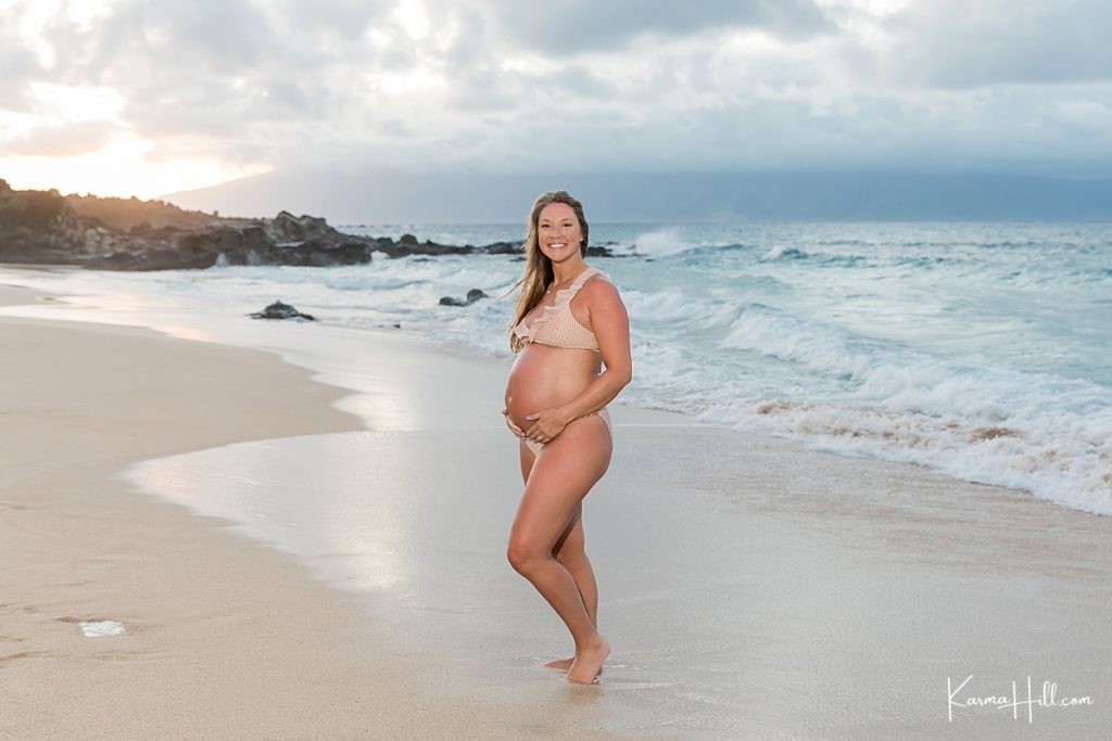 sunset maternity photo with woman in bikini holding pregnant belly on maui beach 
