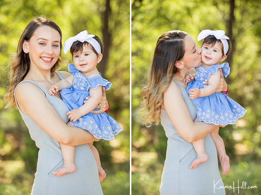 young mother holds toddler daughter with dark hair and white bow and blue dress with trees in the background