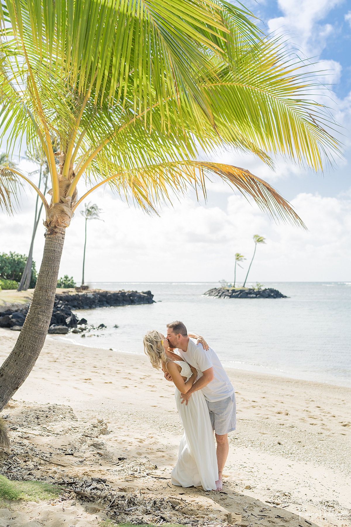 man kisses wife on a hawaii beach with islet in the background and palm trees