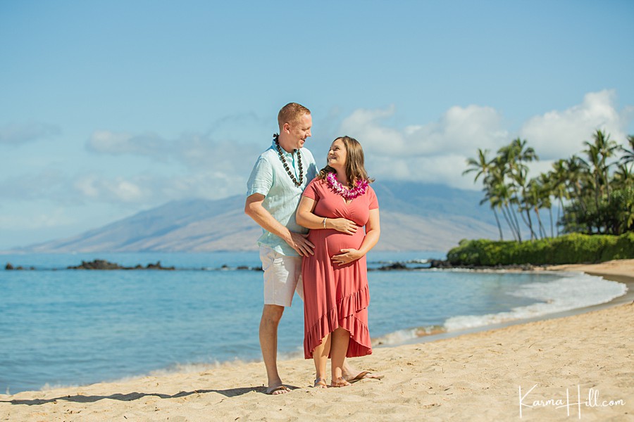 couple stand on the shore of maui with woman holding her pregnant belly 