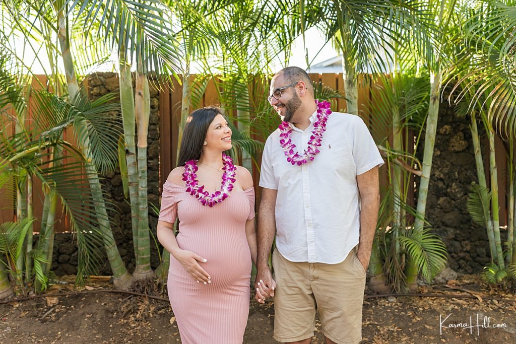 couple wearing leis in front of palm trees with woman holding her pregnant belly 