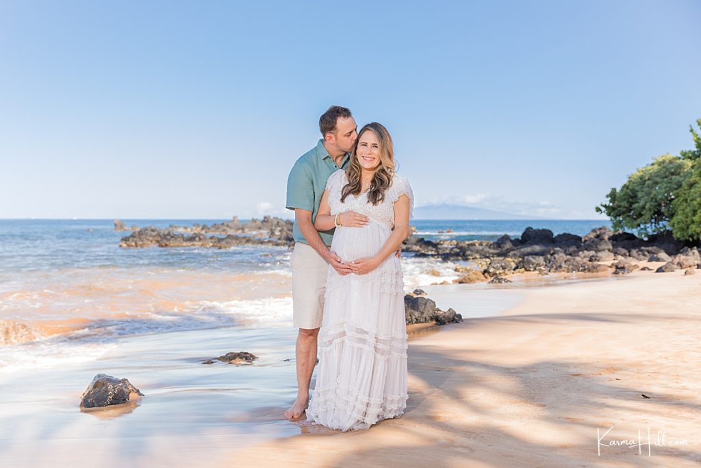 couple embraces and holds woman's pregnant belly in front of the hawaii ocean 