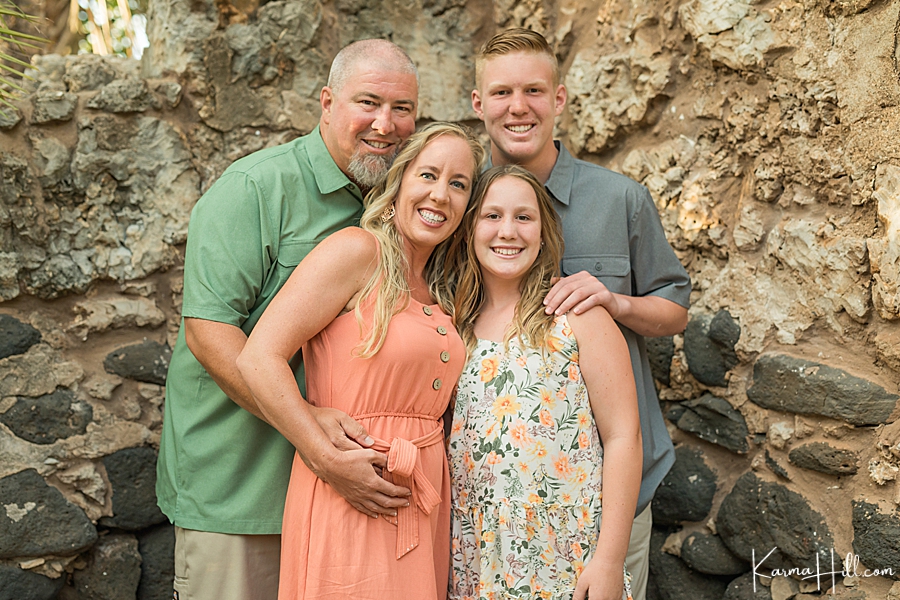 smiling family of four stands in front of rustic rock wall 